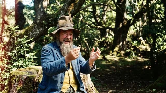 A bearded man in a blue coat and hat laughs joyfully among green foliage, with yellow text asking "Is Tom Almighty?