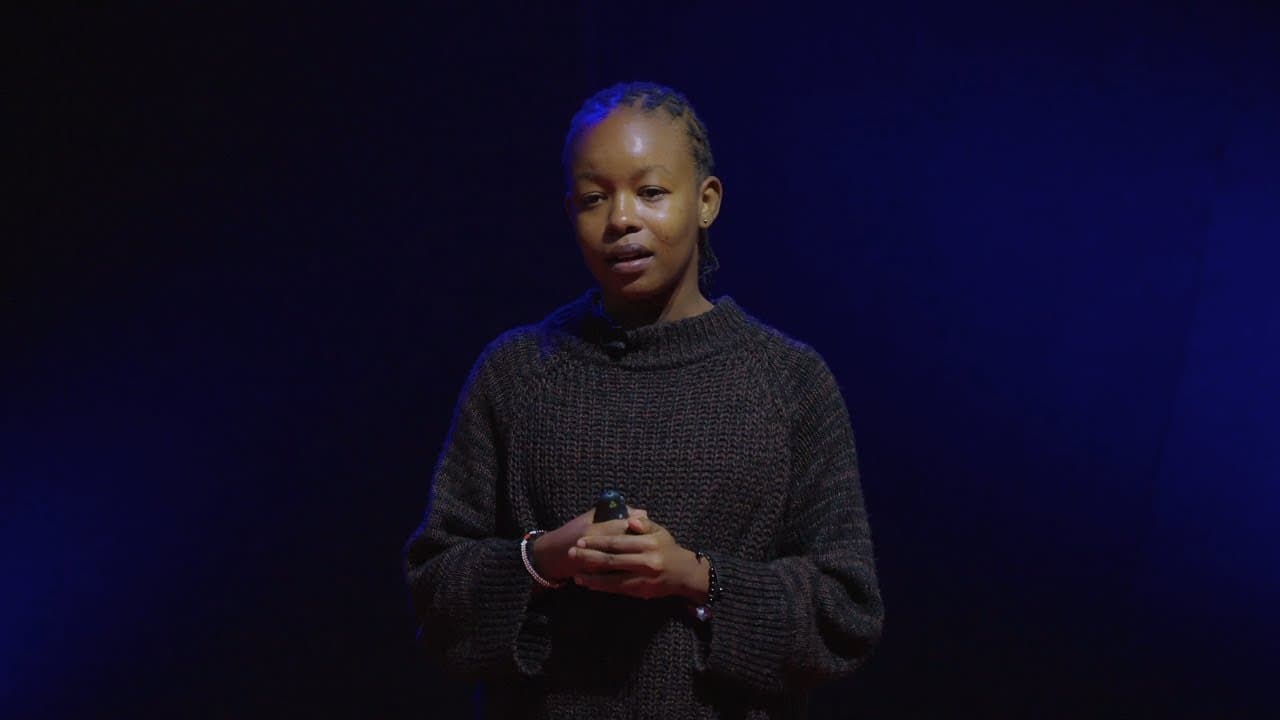 Young woman in dark sweater speaks on stage against blue-lit background, holding microphone during TEDx presentation