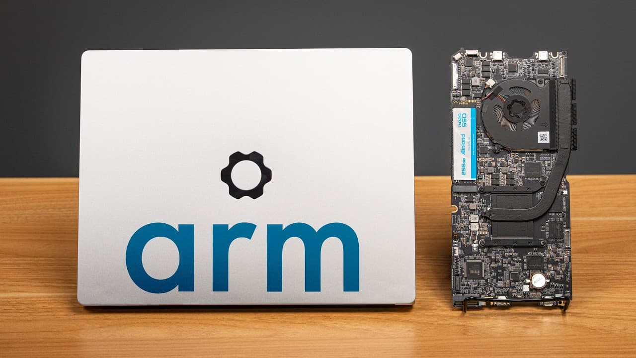 Silver laptop with Arm logo next to exposed computer motherboard with cooling fan on wooden surface