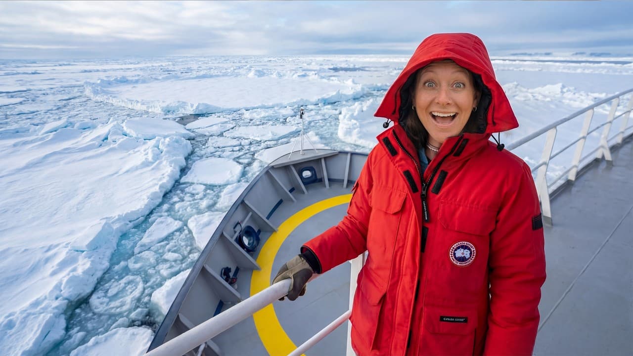 Woman in red parka on icebreaker ship deck smiling at camera with Arctic ice floes and frozen landscape visible behind her