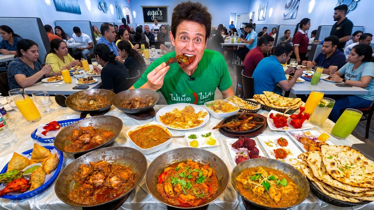 Man in green shirt eating spicy food surrounded by multiple curry dishes at a crowded restaurant with "Ago's" sign visible