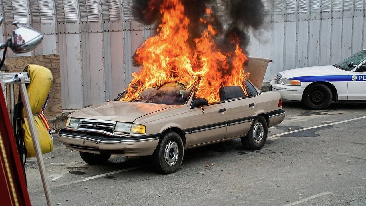 A tan sedan engulfed in intense flames with black smoke billowing upward while a police car and firefighting equipment…