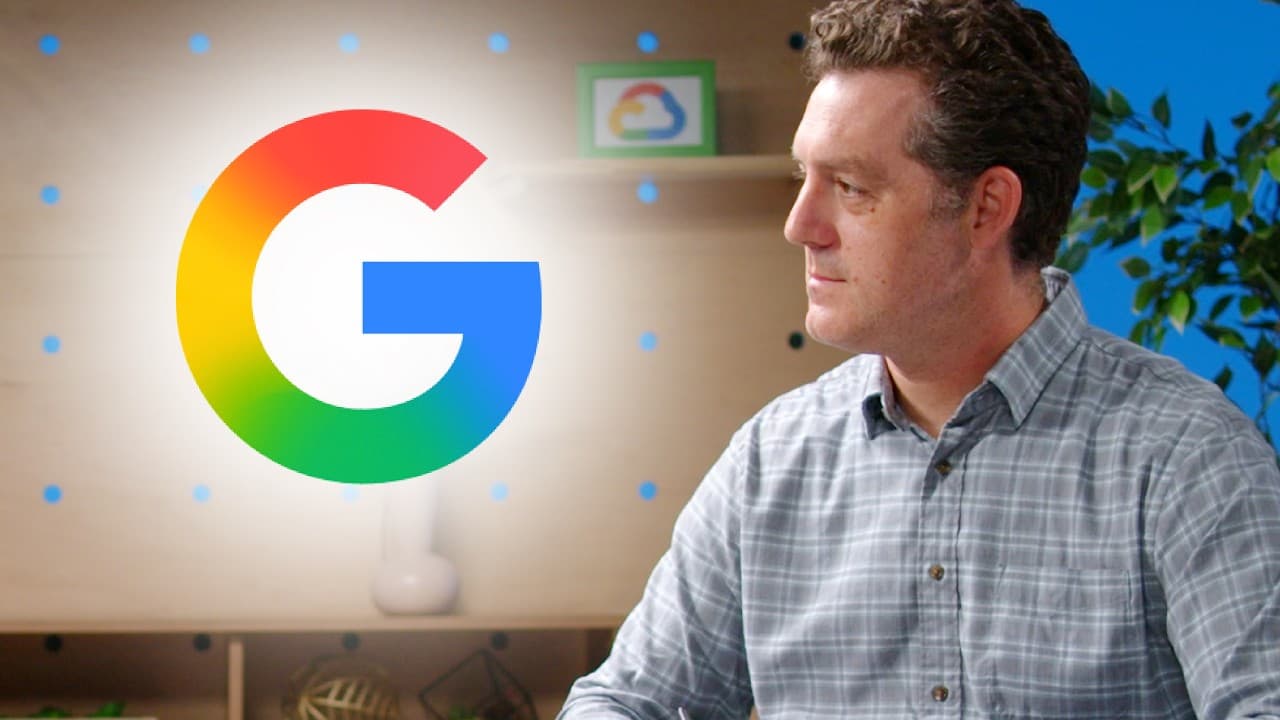 Man in plaid shirt sitting in front of large Google logo in office setting with blue background and plants