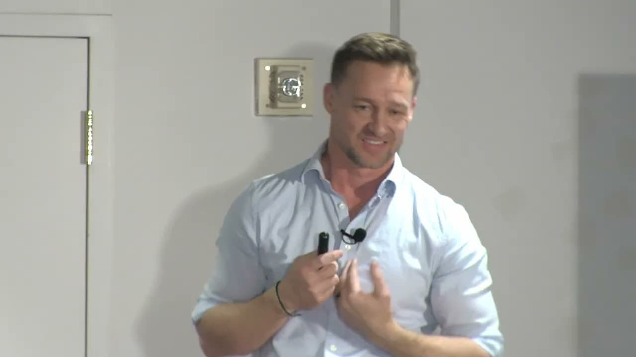 Man in light blue shirt holding microphone while speaking in front of plain wall with electrical outlet visible
