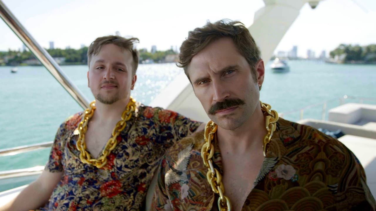 Two men in patterned shirts and gold chains pose on a boat deck with a tropical coastline and water in the background.